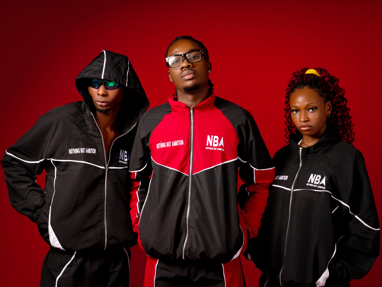 Three models wearing NBA tracksuits and jackets in black and red, on a red studio backdrop