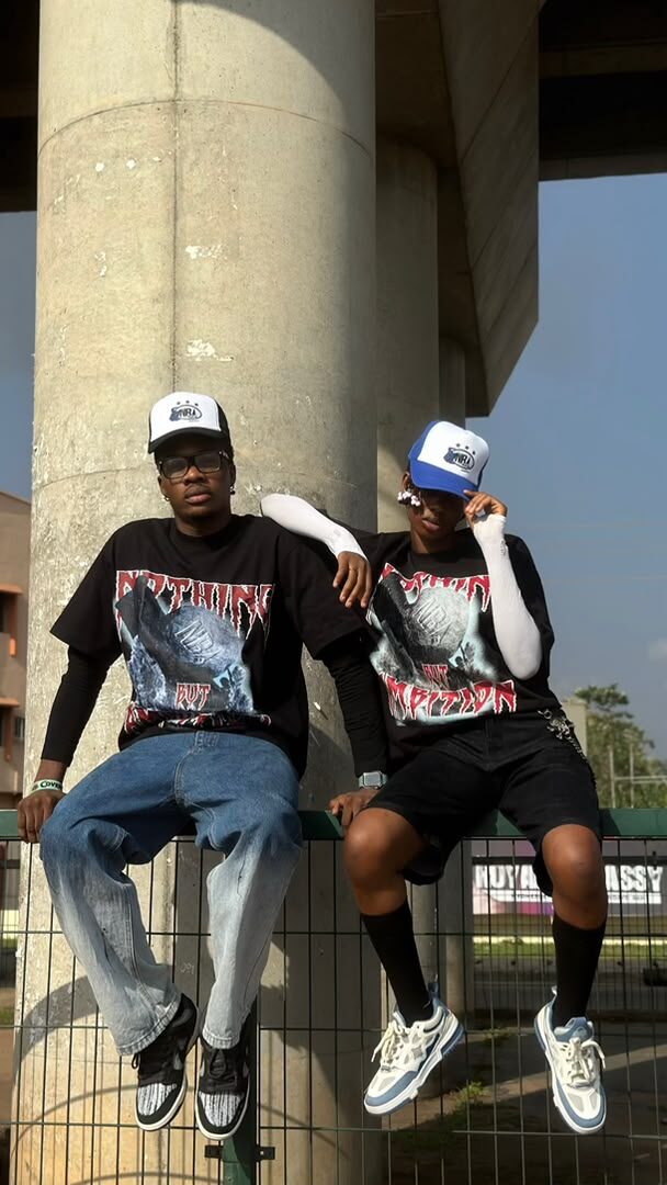 Two models sitting on a fence pillar under a city overpass, wearing NBA graphic tees and trucker caps