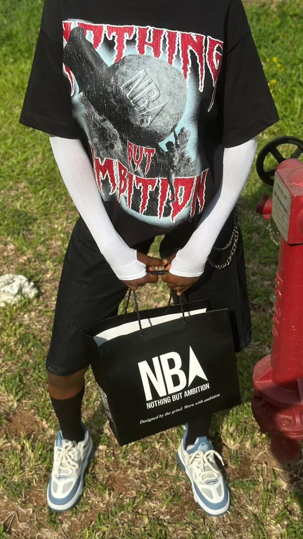 Close-up of a model wearing the black NBA graphic tee, holding an NBA shopping bag, standing by a red fire hydrant