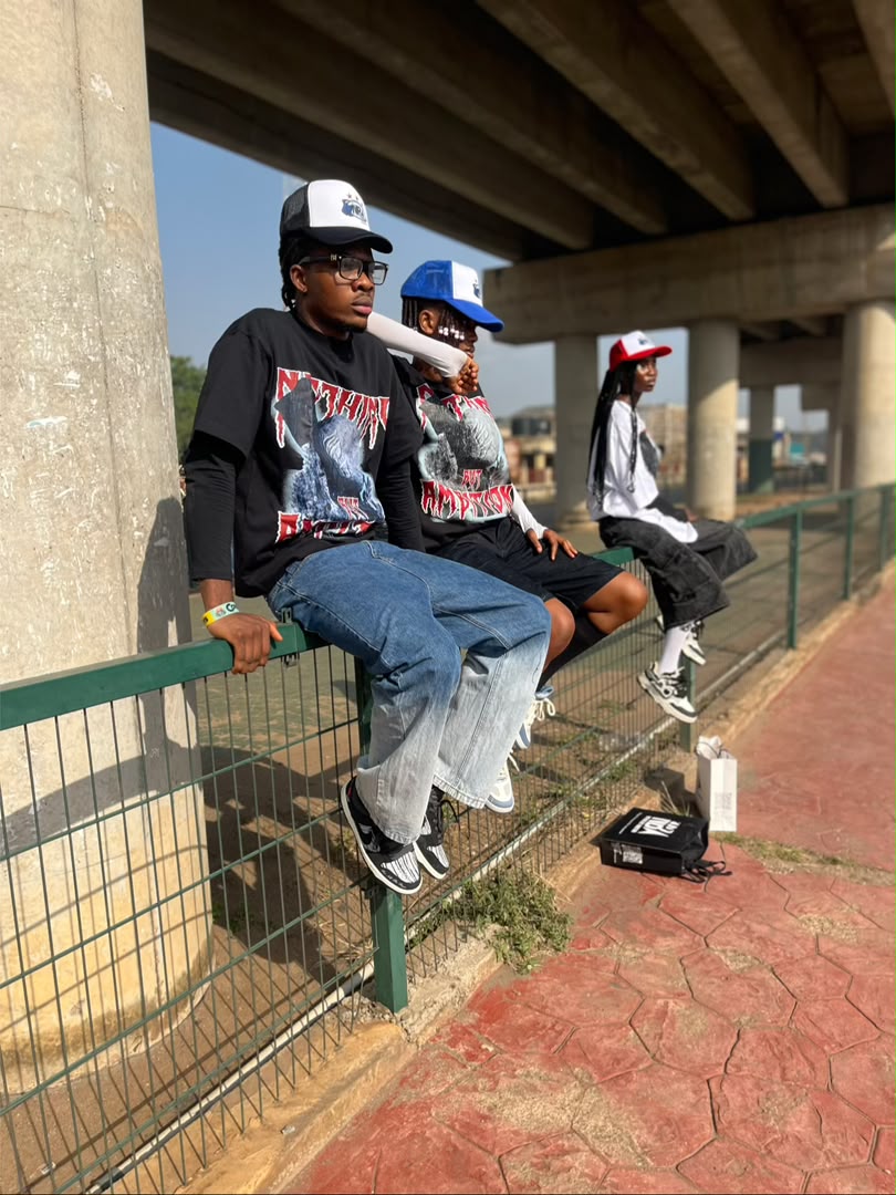 Three models sitting on a green railing under a city overpass, wearing NBA graphic tees and trucker caps in black, blue and red