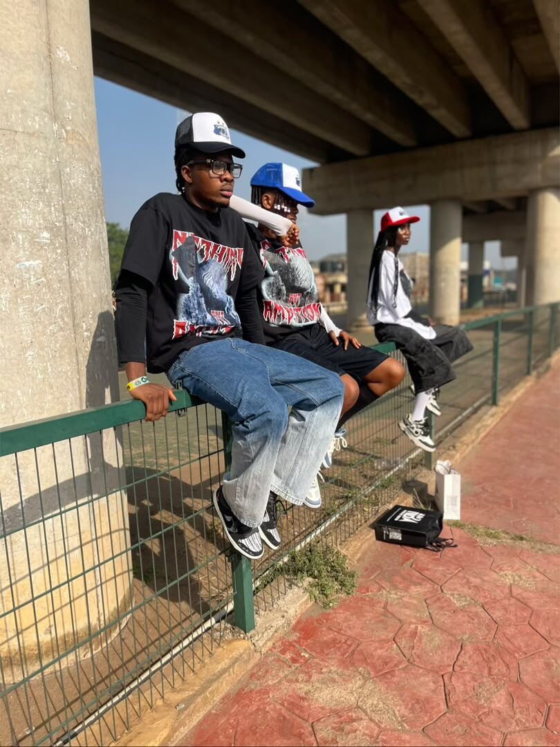 Model crouching in the black NBA graphic tee and red trucker cap, holding an NBA shopping bag, radio tower in the background