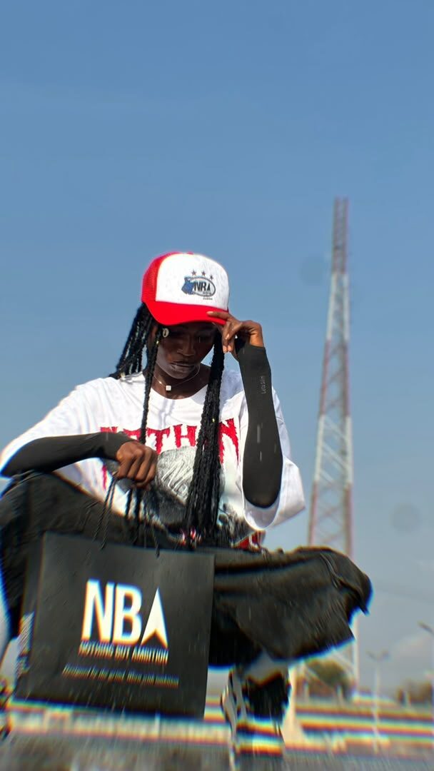 Three models sitting on a green railing under a city overpass, wearing NBA graphic tees and trucker caps