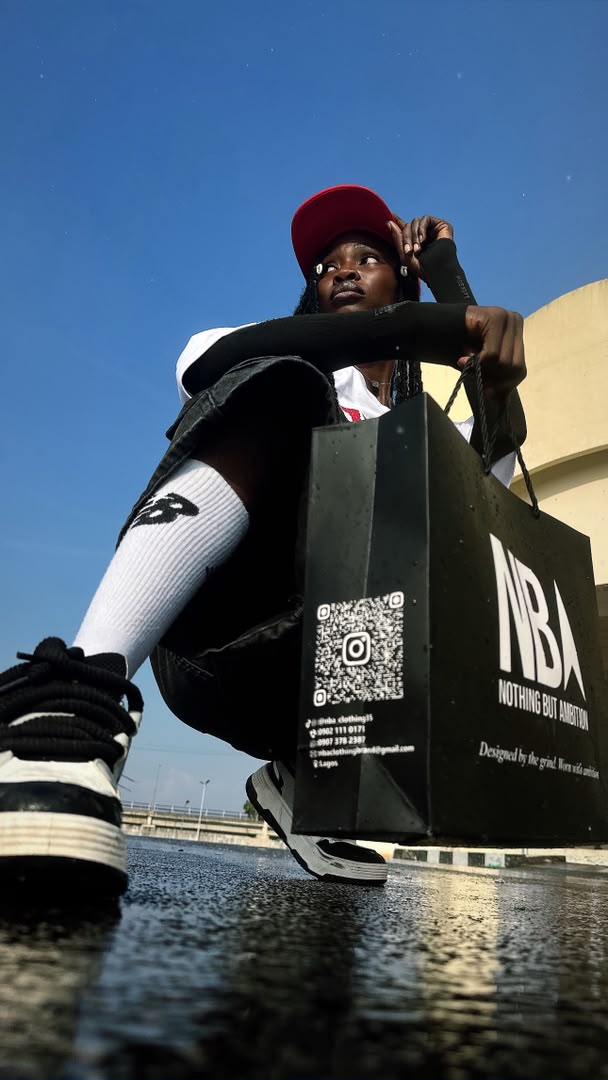 Model crouching low-angle in the black NBA graphic tee and red trucker cap, resting on the NBA shopping bag on wet reflective ground