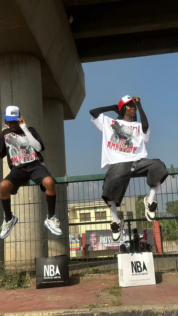 Two models jumping mid-air on a fence under a city overpass, wearing NBA graphic tees in black and white with trucker caps