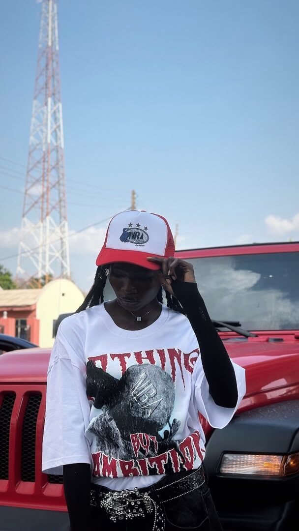 Model wearing the white NBA graphic tee and red trucker cap, adjusting the cap, radio tower and red Jeep in the background