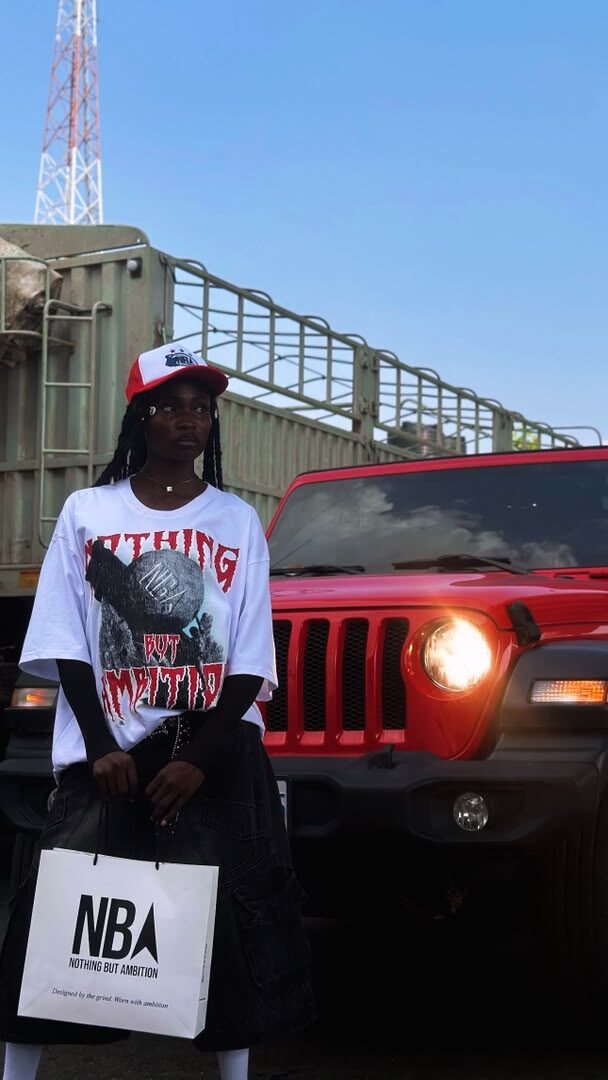 Model wearing the white NBA graphic tee and red trucker cap, standing beside a red Jeep, holding an NBA shopping bag