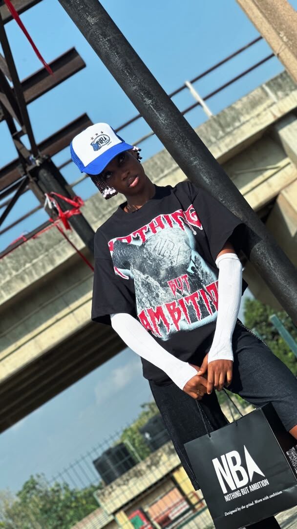 Model wearing the black NBA graphic tee and blue trucker cap, holding an NBA shopping bag, industrial pole in the background