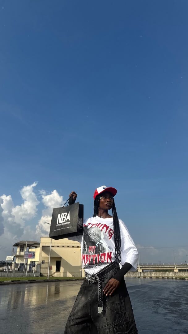 Model wearing the white NBA graphic tee and red trucker cap, holding an NBA shopping bag overhead, wet reflective ground