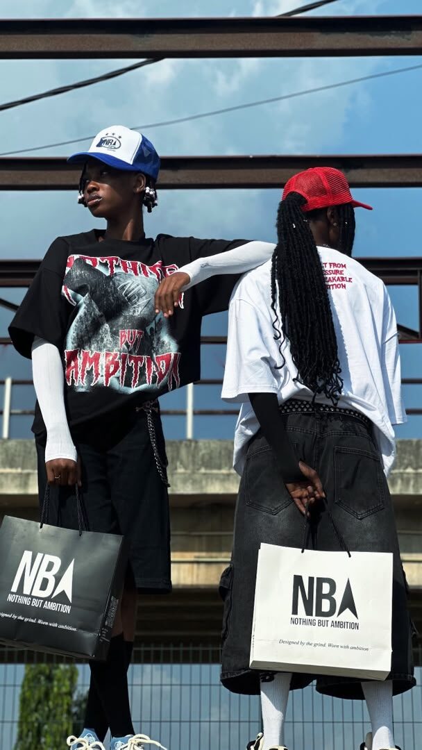 Two models wearing NBA graphic tees in black and white with trucker caps, holding NBA shopping bags, steel beams in the background
