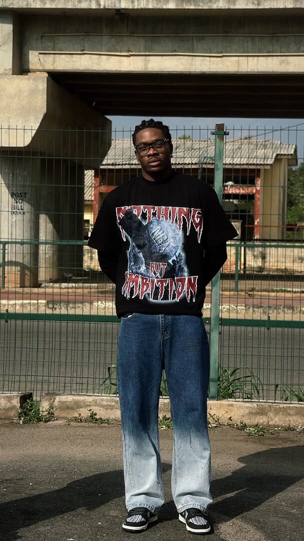 Model wearing the black NBA graphic tee and glasses, standing by a fenced schoolyard