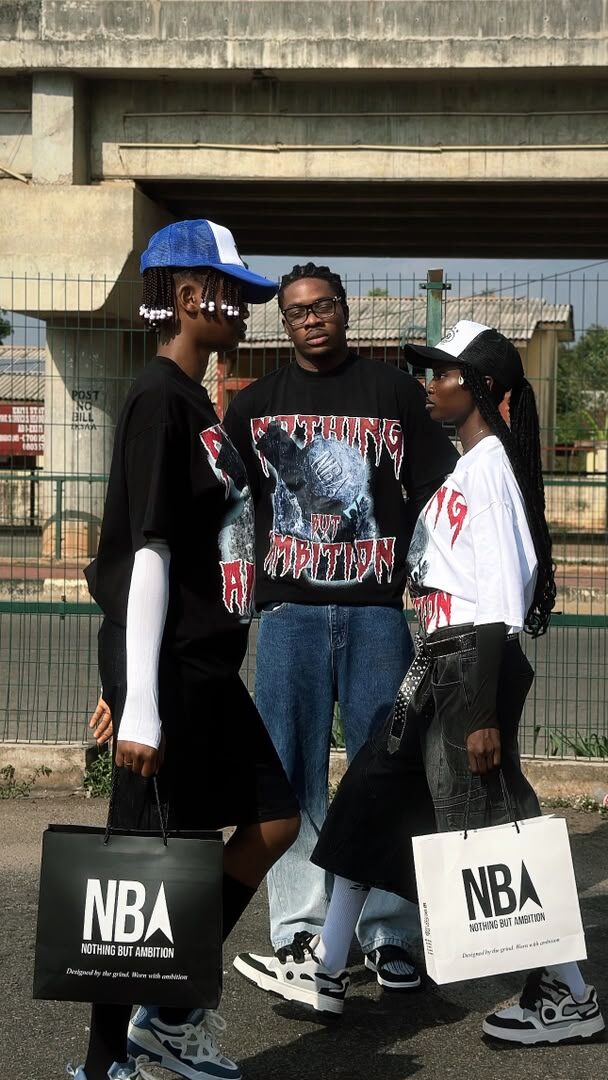 Three models together under a city overpass, wearing NBA graphic tees and trucker caps, holding NBA shopping bags