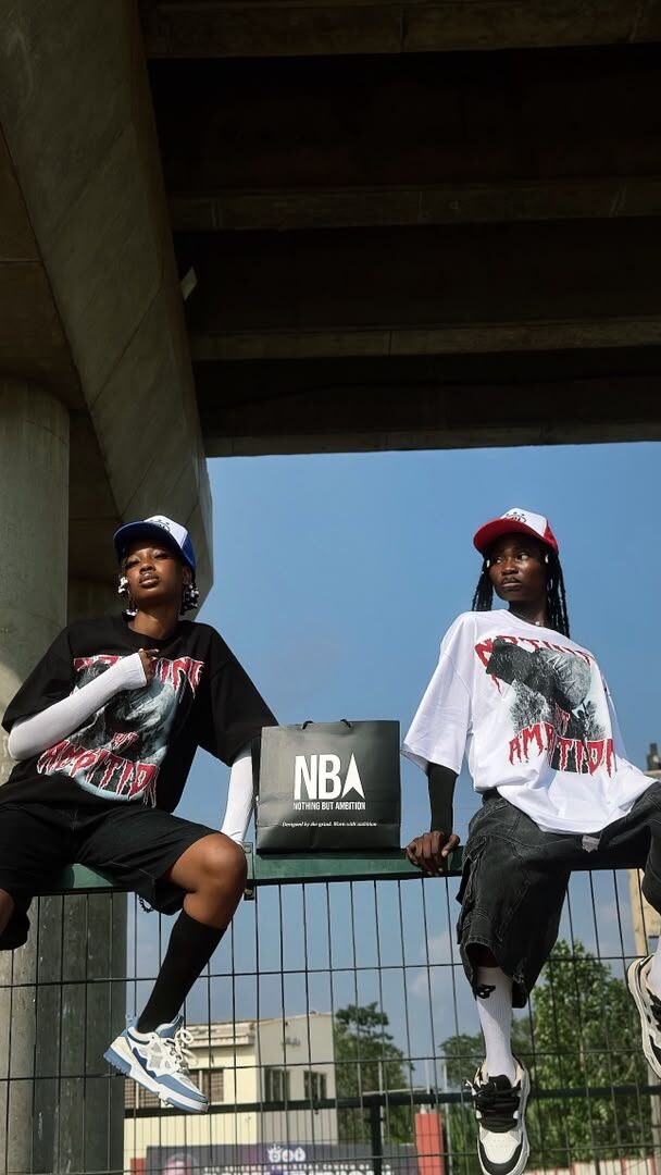 Two models sitting on a railing under a city overpass, wearing NBA graphic tees and trucker caps, with an NBA shopping bag between them